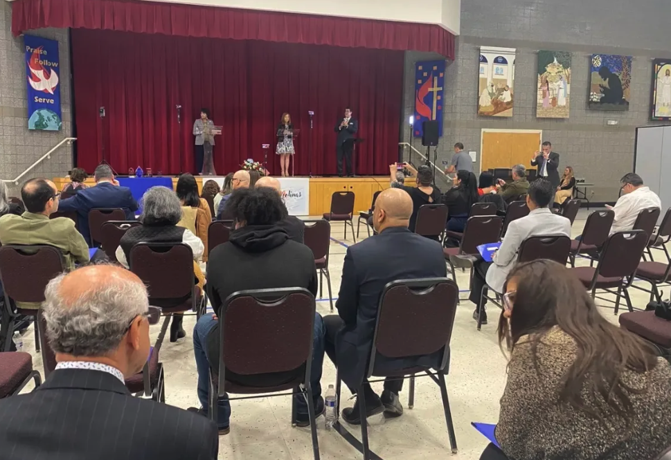 About two dozen people sit a church auditorium and listen to three speakers standing on a stage. The image is shot from behind, in the back of the room. The stage has a red curtain. The walls of the room and decorated with religious banners.